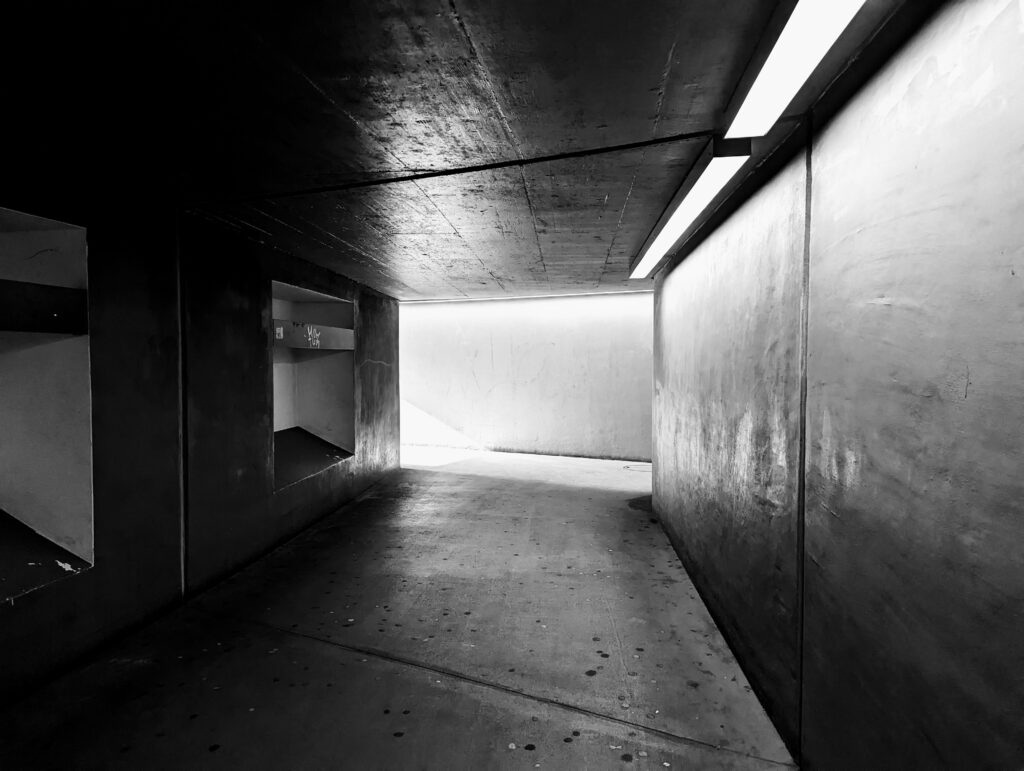 Une photographie en noir et blanc d'un tunnel ou d'un passage souterrain en béton. Le couloir est désert, avec des murs texturés et des niches rectangulaires sur le côté gauche. Une rampe lumineuse au plafond éclaire vivement le mur de droite, menant vers une sortie lumineuse au fond qui crée un fort contraste.